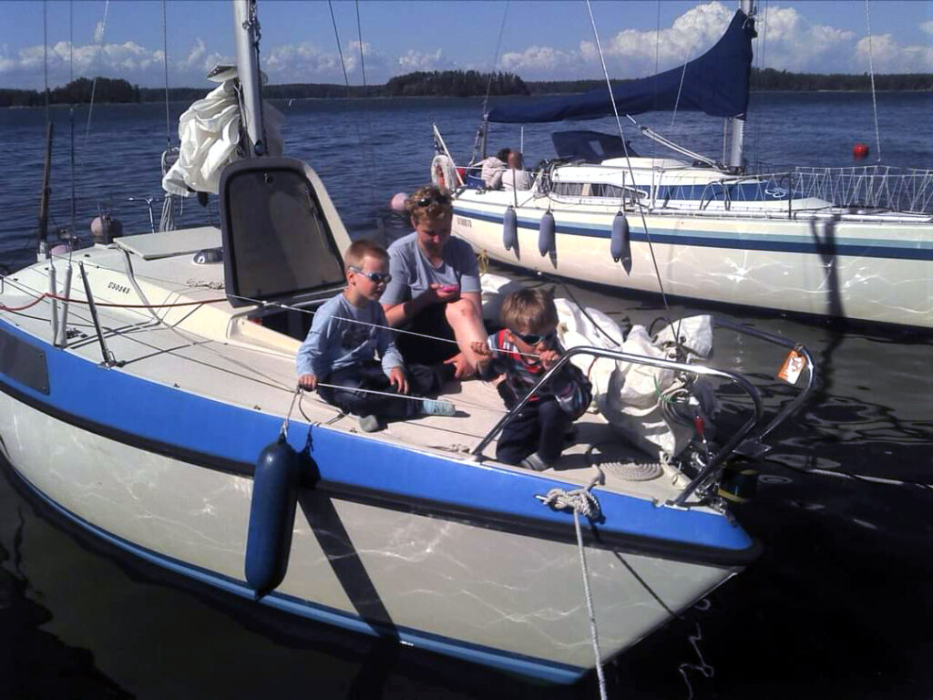 Purjevene laiturissa, nainen ja kaksi lasta istumassa kannella, purjehduksen aloitus meneillään