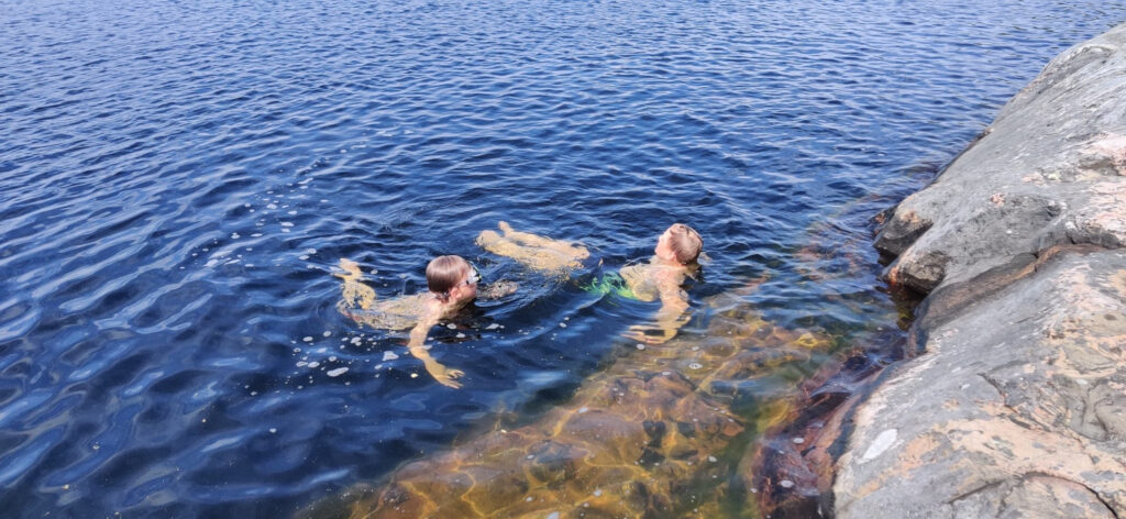 Kallioranta, sininen järvi ja kaksi lasta uimassa