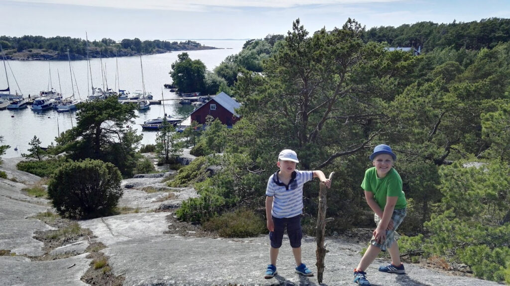 Kaksi lasta kalliolla, taustalla laituri täynnä purjeveneitä.