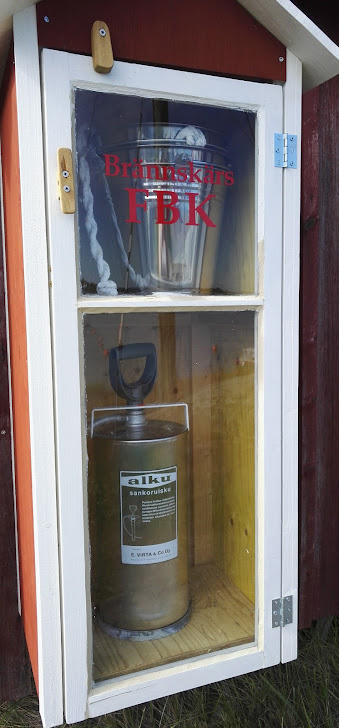 Brännskärin vapaapalokunta: Ämpäri ja pumpattava sammutin lasikaapissa.